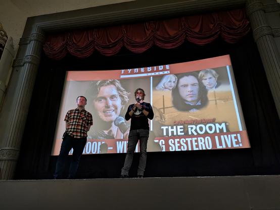 A photo of Greg Sestero standing on a stage in front of a cinema screen holding a microphone. It is an old fashioned UK news reel cinema with deep red curtains, the walls decorated with art deco designs.