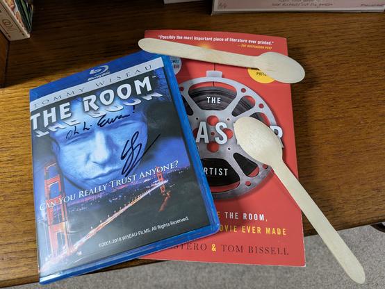 A Blu Ray copy of "The Room" and a copy of Greg Sestero's book "The Disaster Artist" lying on a wooden table together. Two wooden spoons are scattered over them. Both the book and the Blu Ray were sign by Greg with "Oh, hi Ewan!"