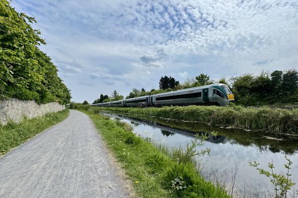A landscape shot of a silver and dark green railcar-type train with a yellow nose and a rounded look approaching along the far bank of a perfectly still canal with a towpath on the left bank with a stone wall on it’s left side. Both sides of the scene are lines with trees and there is a bluer sky with a dappling of very small and thin fluffy clouds overhead.