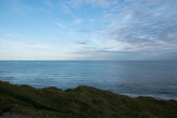 The image shows a scenic view of the coast, capturing the tranquil expanse of the ocean under a sky with scattered clouds. In the foreground, there are grass-covered sand dunes, which add a natural ruggedness to the scene. The overall mood is peaceful and expansive, typical of a coastal landscape.