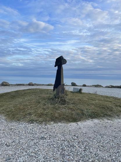 The image features a sculpture set in a coastal environment, likely by a beach or seaside. The sculpture appears abstract, consisting of a dark, textured column topped with a form that might suggest a head or an abstract shape. The ground surrounding the sculpture is covered with small stones and patches of grass. In the background, there is a calm sea and a scattered array of clouds in the sky, contributing to a serene and scenic view. The overall atmosphere is peaceful, enhanced by the natural light and soft color palette.