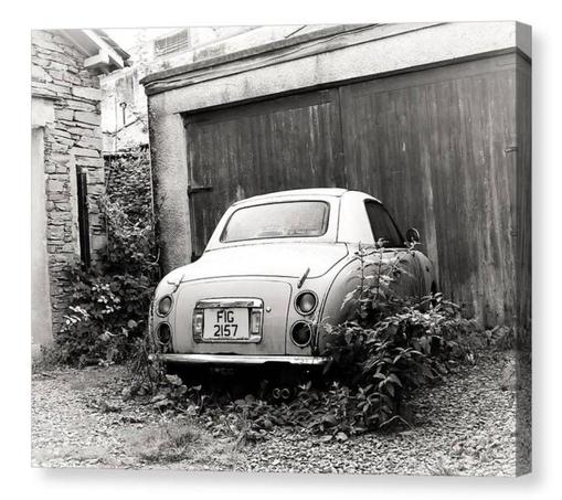 Black and white photograph of a Nissan Figaro car looking neglected.  It's parked up to an old garage and weeds grow up around the rear end.  The image is shown printed upon a block canvas.