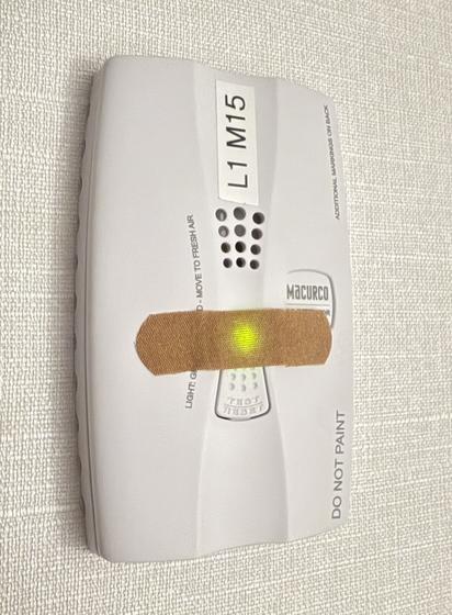 A white plastic co2 or smoke detector on hotel room wall with a brown bandaid placed across a bright lime green LED in the middle which still manages to glow through the layers of fabric.