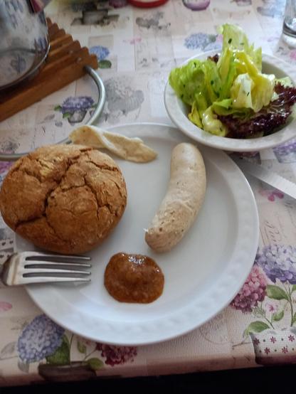 Im Vordergrund ein weißer Teller mit einer Weißwurst, Brötchen und einem Klecks Süßem Senf. Oben rechts ein weißes Schüsselchen mit grünem Salat.