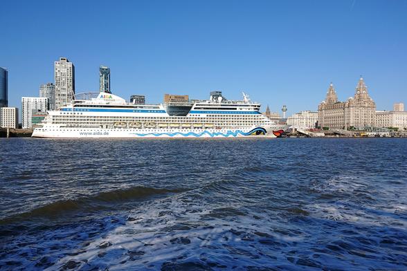 Looking back from the Mersey ferry to the Pier Head.  A huge cruise ship, its hull painted with an eye and huge red lips at the bow, is moored at the cruise terminal; just next to the Liver Building