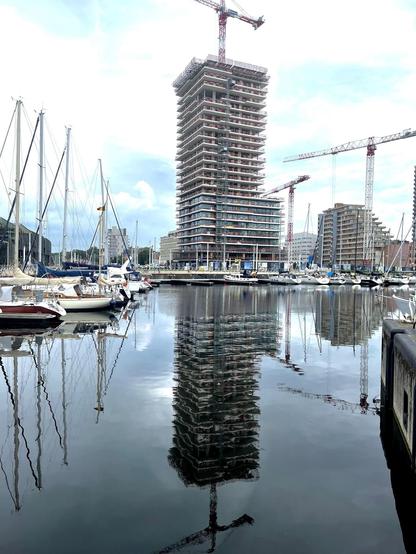 The image features a harbor scene where a large construction project is evident. There's a tall building under construction, characterized by its numerous floors with visible scaffolding and surrounded by cranes. The building is prominently reflected in the calm water of the harbor. Docked in the harbor are several boats, primarily sailboats, adding to the maritime setting of the scene. The environment suggests a blend of urban development and recreational marine activity, likely situated in a city with waterfront properties or developments.