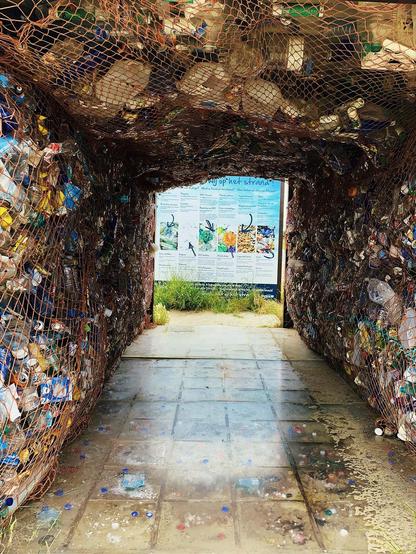 This image shows an arched tunnel-like structure made from netting filled with various pieces of plastic waste and trash, predominantly bottles. It appears to be an environmental artwork or installation meant to raise awareness about plastic pollution. At the end of the tunnel, there's a board with educational materials or information, likely explaining the impact of plastic waste on the environment. The setting looks to be outdoors, as indicated by the daylight and the presence of vegetation at the tunnel's exit.