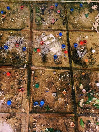 This image shows wet concrete tiles covered in an assortment of small, discarded objects, including colorful bottle caps, can tabs, and bits of broken glass. There are some areas where bubbles have formed, possibly from standing water mixing with some sort of detergent or residue, which suggests this could be an outdoor setting that has been exposed to rain and pollution. The presence of debris and waste gives the scene a neglected, urban feel.