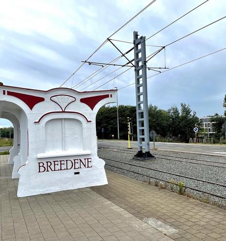 This image shows a tram station named "Bredene." The station has a distinctive architectural style featuring white walls with red detailing. The structure appears to be part of the station design, likely for aesthetic or shelter purposes. There are multiple railway tracks visible, along with electrical lines above for the tram system. The backdrop includes a view of a road and some greenery, hinting at a suburban or semi-urban environment.