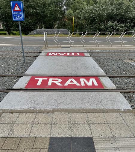 The image shows a tramway crossing integrated with pedestrian pathways. There's a road sign saying "VOORRANG TRAM," which translates to "Priority to Tram" in English, indicating that trams have the right of way at this crossing. The ground features text "TRAM" painted in red on a white concrete surface, likely to alert road users of the tram tracks. The pavement includes textured tiles for safety and accessibility, likely aiding visually impaired pedestrians. Additionally, there are metal railings on one side, probably to guide pedestrian movement and ensure safety near the tram tracks.