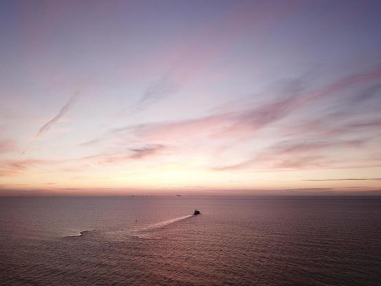 This image captures a serene seascape at sunset. The sky displays a beautiful gradient of colors from soft pink to golden hues, with wispy clouds adding texture to the sky. Lower in the scene, the calm ocean is punctuated by a lone boat moving towards the horizon, leaving a trail in the water. The sea stretches out widely, meeting the sky at a distant horizon. This scene conveys a sense of tranquility and vastness, highlighting the beauty of nature during a sunset over the ocean.