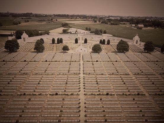This image shows a large military cemetery, evident from the orderly rows of grave markers. In the center, there appears to be a monument or a memorial chapel, surrounded by landscaped grounds including paths and trees. The cemetery is expansive, with a vast number of graves stretching out towards the horizon. The sepia tone of the photo adds a somber, historical feel to the scene. This type of cemetery is generally dedicated to soldiers who have died in wars, serving as a place of remembrance and honor.