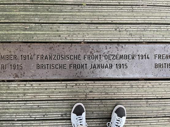 The image shows a metal plaque embedded in a wooden boardwalk. The inscription on the plaque is in German and English and refers to historical military fronts from World War I. It mentions the "Französische Front Dezember 1914" (French Front December 1914) and the "Britische Front Januar 1915" (British Front January 1915). At the bottom of the image, a pair of shoes can be seen, suggesting that someone is standing and looking down at the plaque. This type of installation is likely part of a historical site or memorial walking path dedicated to remembering and providing information about World War I.