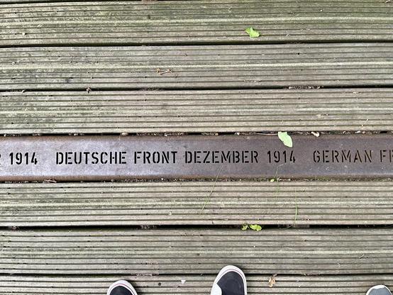 The image shows a close-up view of a wooden boardwalk with a metal strip inset between the planks. The metal strip is engraved with text that reads "1914 DEUTSCHE FRONT DEZEMBER 1914 GERMAN FRONT." This likely commemorates a historical event or location related to a German front line in December 1914, perhaps during World War I. There are also the toes of someone's shoes visible at the bottom edge, indicating they are standing on the boardwalk. A few leaves and some debris are scattered on the wooden surface.