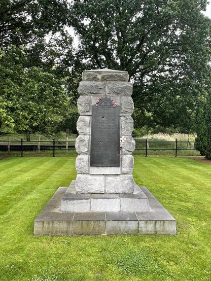 The image shows a stone memorial monument, likely a war memorial, with a large plaque at the front. The plaque features inscriptions and a crest at the top, indicating the memorial may be dedicated to specific individuals or a group from a military unit or community. The structure is surrounded by a well-maintained grassy area, with tall trees in the background, and a metal fence visible in the distance. Red commemorative poppies, symbols of remembrance and respect for fallen soldiers, adorn the plaque.