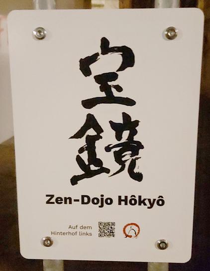 Ein weißes, hochrechteckiges Schild mit schwarzer japanischer Kalligraphie im Mittelpunkt (zwei Schriftzeichen untereinander). 
Darunter steht

Zen-Dojo Hokyo, darunter eine Wegbeschreibung ein QR-Code und das Logo der Zen-Vereinigung.