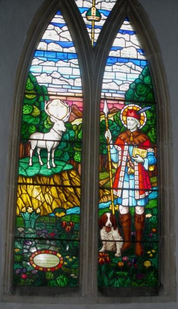 Photo of a two-panel stained glass window from St Ethelbert's Church in Herringswell, Suffolk, UK.

It depicts St Hubert. In the right panel, a medieval dressed hunter stands with a spear in one hand and a book in the other. Around his head is the glow of a halo. Near his feet is a small brown and white dog. In the left panel, a stag is standing on a hill. Between its large horns is a glowing Christian cross. 

St. Hubert's story holds that after the death of his wife, the nobleman went to live in the woods. There, he saw a stag with a cross glowing between its horns, and took it as a sign from God. 

Window in memory of Llewellyn Sidney Davies. Designed by H.W.Luxford.