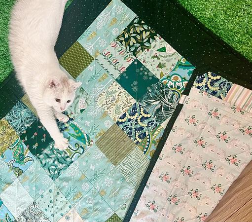 White cat stretched out on her side, one paw with claws slightly out in the top left corner of a patchwork quilt, with a bit of white cat fabric showing. Cat is very cute.