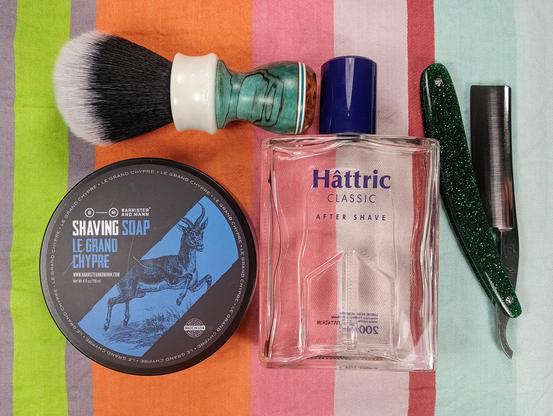 (Le Chat Mistral description)

The image showcases a collection of grooming items arranged on a striped background. At the top left, there's a shaving brush with a white handle. Below it, there's a round tin of shaving soap labelled "Le Grand Chypre" with an image of a goat. To the right of the shaving soap, there's a clear bottle of aftershave labelled "Hâttric Classic." Further right, there's a straight razor with a green glittery handle. The items are neatly laid out, suggesting a traditional shaving setup.