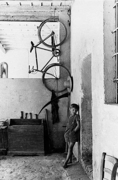 un jeune garçon désoeuvré appuyé contre un mur, les mains dans les poches, à côté de son vélo suspendu au plafond de l’atelier