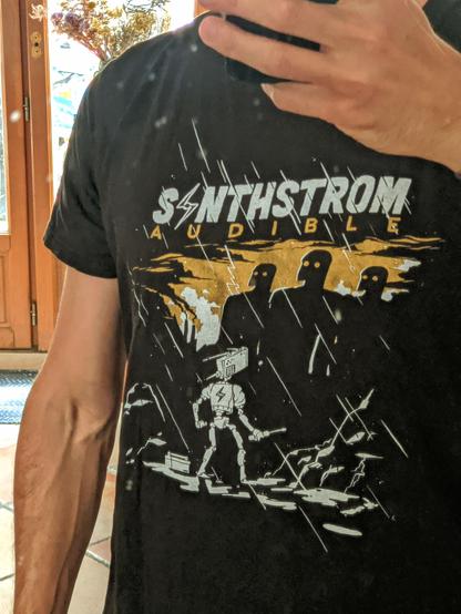 A headless selfie. It shows a black shirt with a drawing on it: it's a small cute robot facing three big corporate robots - aka Synthstrom against Big Synth. It's raining, obviously, because Synthstrom is the maker of the Deluge groovebox.