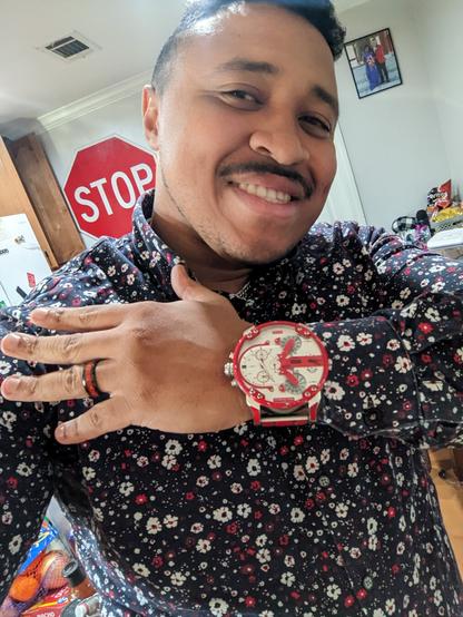 Selfie of a black man smiling while displaying a very large red and white watch and a black button up shirt patterned with white and red flowers