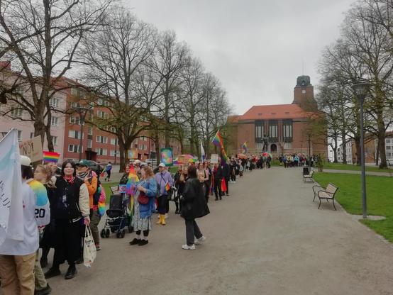 Pride-kulkue on lähdössä liikkeelle Lahden kaupungintalon edustalta. Kaupungintalo, suuri punainen tiilirakennus jossa on torni, näkyy mäen päällä. Edessä on puistoa, puita ja leveä hiekkakäytävä. Ihmiset seisovat leveässä jonossa lippuineen ja kyktteineen. Taivas on pilvinen.