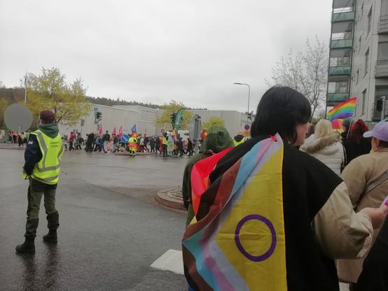 Kulkue ylittää Saimaankatua ja kulkee pääkirjaston ohi, joka on matala valkoinen rakennus edessä. Kirjaston lipputangoissa on pride-liput, tosin ne eivät erotu hyvin kuvassa. Liikenteenohjaaja keltaisessa liivissä pysäyttää autoliikennettä.