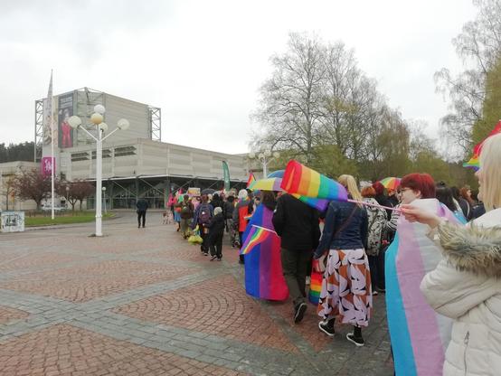 Kulkue saapuu kaupunginteatterin pihaan. Rakennus on harmaata betonia. Ihmisillä on pride-lippuja, jotkut pitävät niitä viittoina.