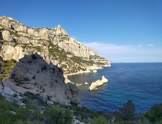 Vue sur la candelle, après avoir passé l'échelle (oui c'est une vraie échelle) de la plage de Sugiton.

Au premier plan un petit rocher et de la végétation, puis on voit la mer avec le "torpilleur", une petite île calcaire en forme de sous-marin.

Sur la gauche la côte formée par les calanques et son calcaire blanc. Les falaises de la grande candelle (entre coupée par une large vire herbeuse)

Au loin sur la côte on aperçoit le bec de l'aigle (une pointe avec une roche beaucoup plus orange du côté de la Ciotat).

L'horizon est dégagé avec quelques nuages très légers. Autrement le ciel est complètement bleu.