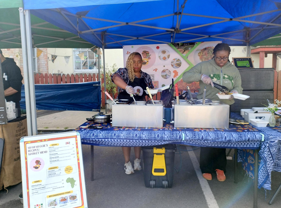 Mami Maggie's Recipes' stall at Roath Market. Two vendors are preparing an order for the customer in front of us, taking servings from two multisection steel food warmers on the table in front of them. The table is covered in a blue printed cloth, the awning is blue too. A menu is placed on the ground to the front left of the stall.