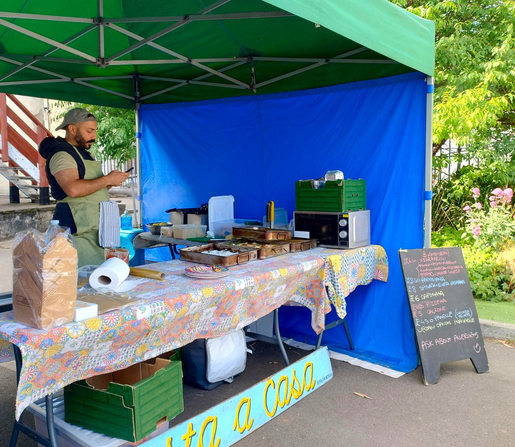 Pasta a Casa's stall at Roath Market. Fabritzio stands behind a table with a bright multicoloured cover. The table is covered with dishes of food, cooking kit and serving items. The stall's awning is green and its side wall is blue. A menu sits on the ground to the right of the stall.