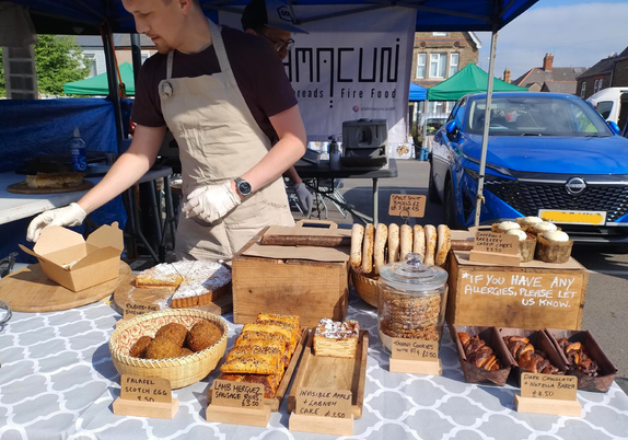 Lahmacun's stall at Roath Market. The vendor is putting part of our order into a card box for us to carry home. A variety of baked goods sits on a grey and white patterned table cover. There are gaps because they're still setting up their display. Spinach and feta borek and date and pecan focaccia had not yet put in an appearance.