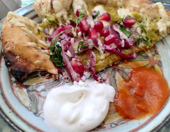 A quarter of Lahmacun's falafel flatbread lies on a craft potter plate, with a part-eaten piece of crust to the left. The flatbread is topped with a layer of crushed falafel, overlaid with drizzled tahini, bright rose-coloured pomegranate seeds and pickled red onions, plus some green herbs. To the front of the image, spoonsful of white garlic and red scotch bonnet sauces.