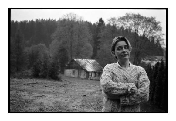 A black and white film photograph showing a woman with her arms crossed, standing outdoors. In the background, there's a rustic building and a forest
