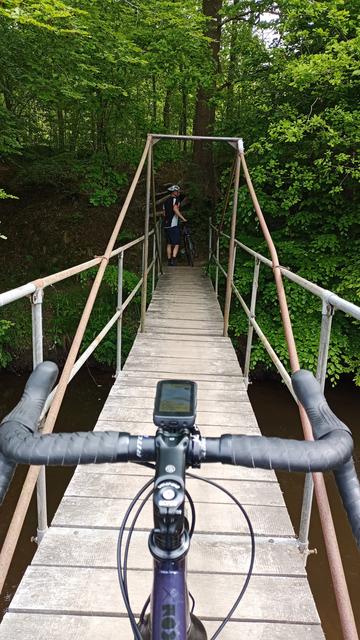 Eine schmale kleine Holzbrücke über einen schmale Fluss. Im Vordergrund ein Dropbarlenker, voraus auf dem anderen Ufer ein Gravelbikefahrer.