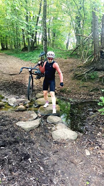 Ein Gravelbikefahrer hat ein Gravelbike geschultert und trägt dieses über Trittsteine in einem fast ausgetrockneten Bach.