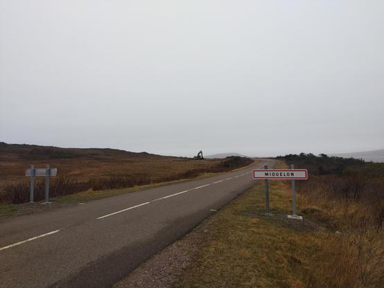 Un paysage de tourbières. Une route part du coin en bas à gauche et traverse l'image. Un panneau indique que l'on rentre dans le village de Miquelon. Au loin, sur une petite crête, on voit une pelle mécanique.