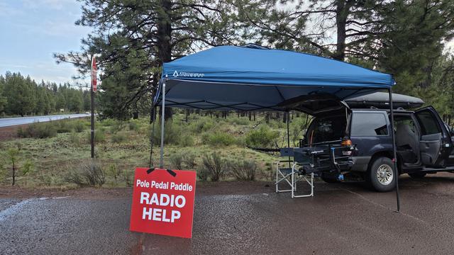 A red sign reads "Pole Pedal Paddle Radio Help". It is taped to a blue-green canopy easy up.