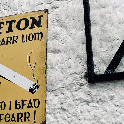 A closely cropped photo shows part of an old cigarette poster in Irish on the left and a black grating/gate on the right against a rough stuccoed off-white exterior wall.