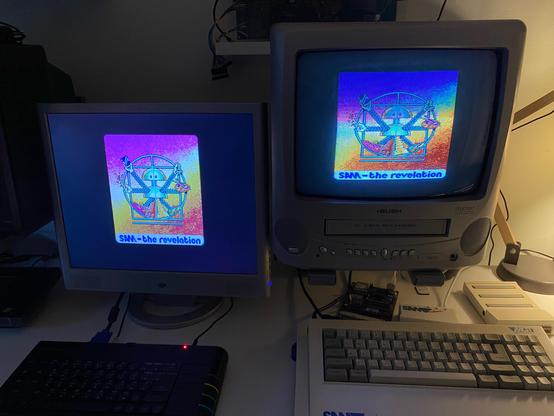 Photograph of two computers and two screen. On the left, ZX Spectrum Next with a flatscreen monitor. On the right SAM Coupé with a portable television set. Both screens show the same “Vitruvian SAM” image.