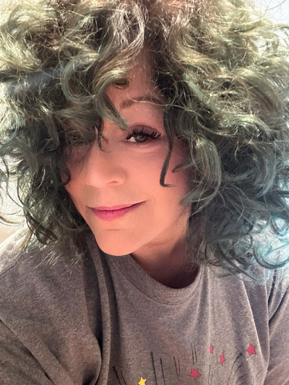 Selfie with smirky face and v wind tousled curly brown hair that happens to be highlighted pale teal green