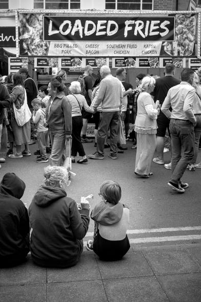 A black and white street scene. The main focus is a woman and two boys in the foreground bottom left. They are sat on the curb of the street with their backs to the camera. The woman in the middle is sharing food on her fork with the boy to her right. Crowds of people are walking in the street. On the other side of the street is a food stand with a sign reading "Loaded Fries". Perhaps the woman is sharing loaded fries with her sons.