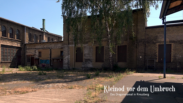 Die Stallungen (gelbe Backsteingebäude davor eine Birke) von hinten, vorm Dorfplatz aus gesehen