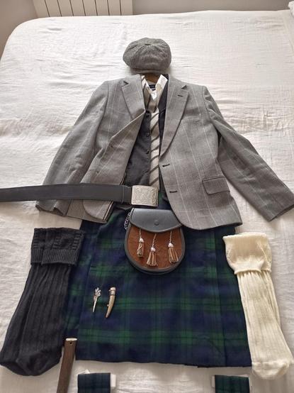 Atuendo sobre cama. Colcha de comer crudo y sobre ella, colocados en su posición una vez puestas, las siguientes prendas: gorra newspaper gris tweed espina de arenque, camisa blanca, chaleco gris oscuro espina de arenque, chaqueta de cuadros príncipe de gales, corbata de lana en líneas gruesas gris claro y gris oscuro, kilt black watch, sporran con frontal de pelo corto marrón, un cinturón de hebilla grande plateada con un dragón céltico, un par de medias negras a un lado y otras color crudo al otro y borlas black watch. 
En el kilt, dos alfileres, uno plateado con un ramito de cardos escoceses y otro hecho de una punta de cuerno de ciervo.