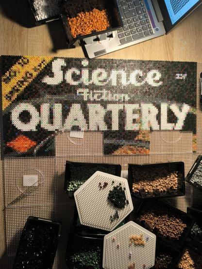 View of my recent artwork: on top there are the words "Science Fiction Quarterly" with a black and grey background. On the top left corner is a yellow stripe with the words "No. 10 Spring" but it's very pixelated. On the remaining pegboards that are not yet in use, there are small boxes with ironing beads in different colors.