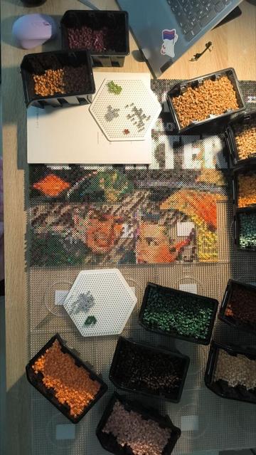 View of my recent artwork: two faces, not yet fully filled in. On the left is a man with a hat, an officer, and on the right is a woman, looking at him. On the remaining pegboards that are not yet in use, there are small boxes with ironing beads in different colors.