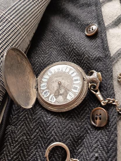 Primer plano, reloj de bolsillo sobre chaleco gris oscuro en espina de arenque. La esfera tiene el fondo grabado en plata oscura con motivos vegetales y números romanos en negro y enmarcados con paréntesis plateados sobre fondo blanco. Tiene un segundero pequeño en la mitad inferior y las manillas en negro también en motivos vegetales.