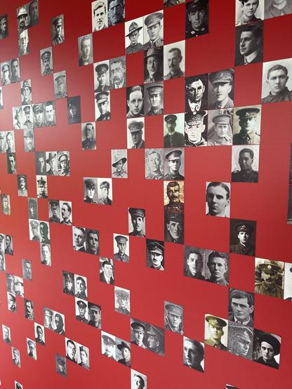 This image shows a collection of black and white photographs arranged on a red wall. Each photograph portrays an individual, predominantly males, most likely soldiers or military personnel given that several are wearing uniforms featuring caps or hats typical of military attire. The style of uniforms and the photographic quality suggest that these images could be historical, possibly from the early to mid-20th century. The red background creates a striking contrast with the black and white photos, emphasizing each individual portrait. This display could be part of a historical exhibit, memorial, or a tribute to military personnel from a specific period.