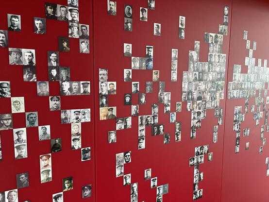 This image shows a large number of black and white photographs displayed on a red wall. The photographs appear to be arranged in clusters or blocks across the wall, and they depict various individuals. Such a display could be part of an exhibition, memorial, or gallery intended to present historical figures, commemorate certain individuals, or illustrate a particular era or theme. The setup creates an impactful visual tribute or historical representation.